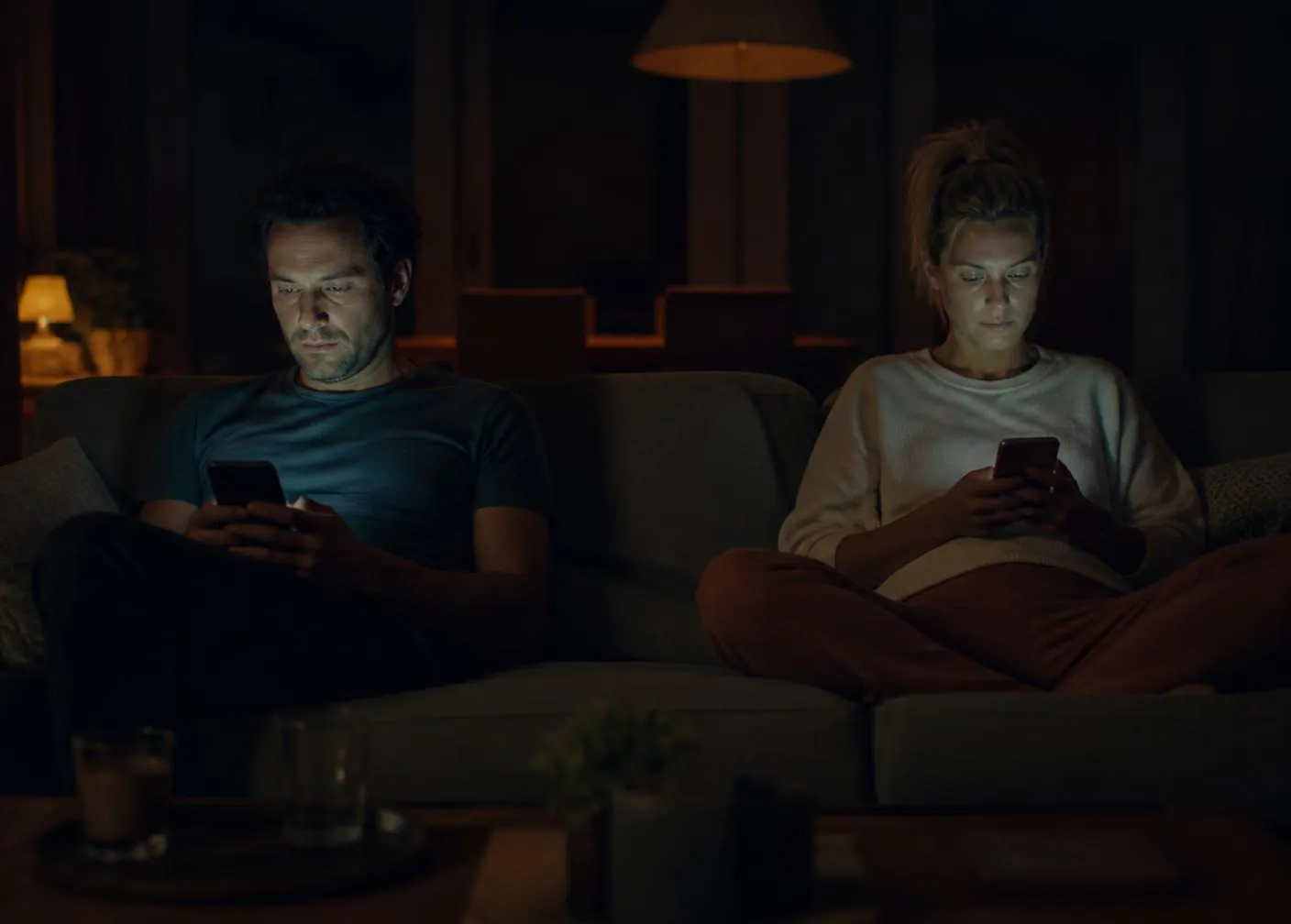 Casal sentado no mesmo sofá, cada um olhando para o próprio celular em um ambiente escuro